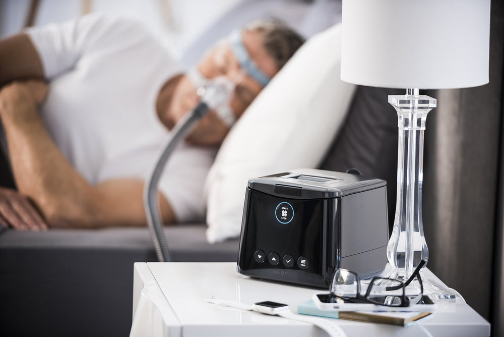 SleepStyle Auto CPAP device sitting on nightstand with Man sleeping wearing sleep mask