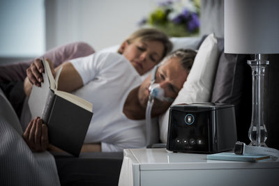 SleepStyle Auto CPAP device sitting on nightstand with Man reading in bed wearing sleep mask